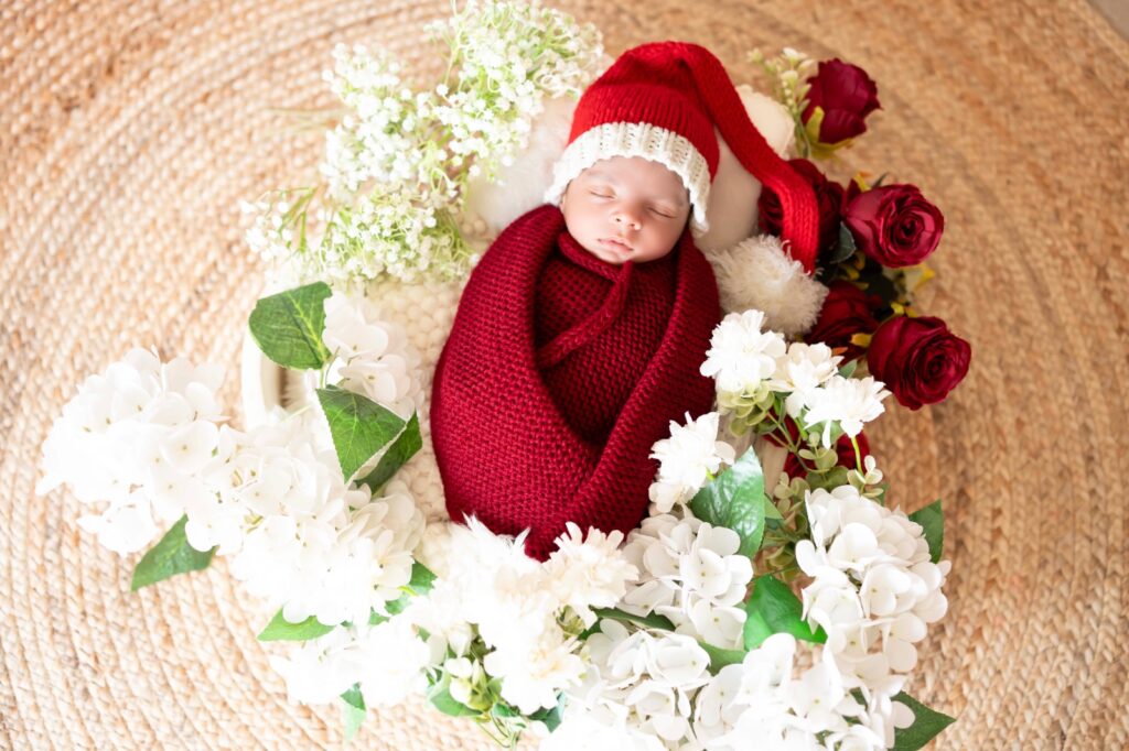 Baby wrapped in beautiful red wrap and setup made with white flowers wearing red cap, photography done by one of best photographer in Gurgaon