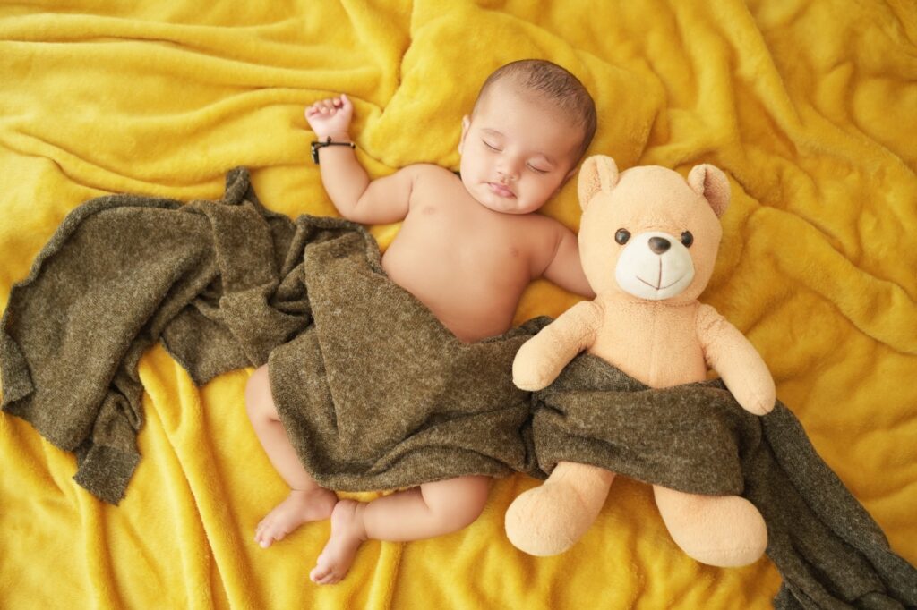 Baby sleeping on yellow velvet background with teddy in his hands and photography done by one of best photographer in Gurgaon